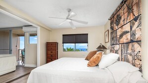 A bedroom with a bed, a ceiling fan, a window with a view, a wooden cabinet, and a door leading to a balcony. - Kailua Village#604 (Kailua-Kona)
