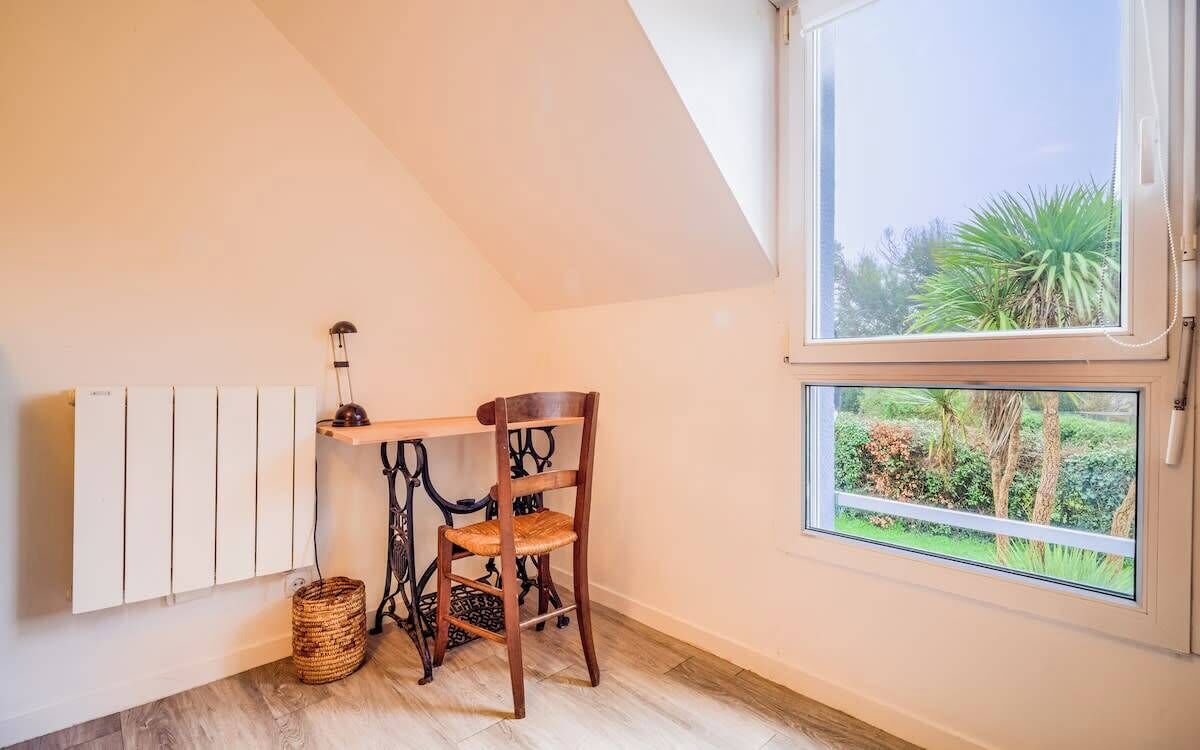 A small, cozy room with a sloped ceiling, a radiator, a wooden chair, a small table, and a window with a view of greenery.