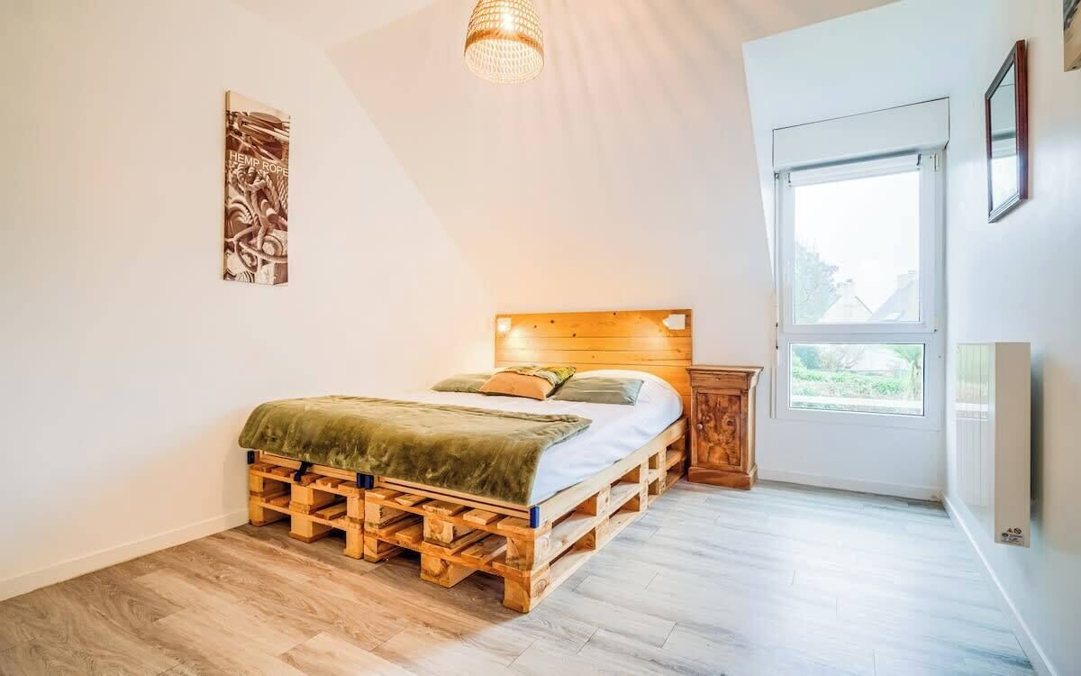 A bedroom with a bed made of wooden pallets, a wooden headboard, a small wooden nightstand, and a window with a view of greenery.