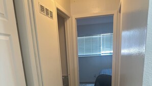 Un pasillo estrecho con suelo de madera que conduce a un dormitorio con cama y ventana con persianas.