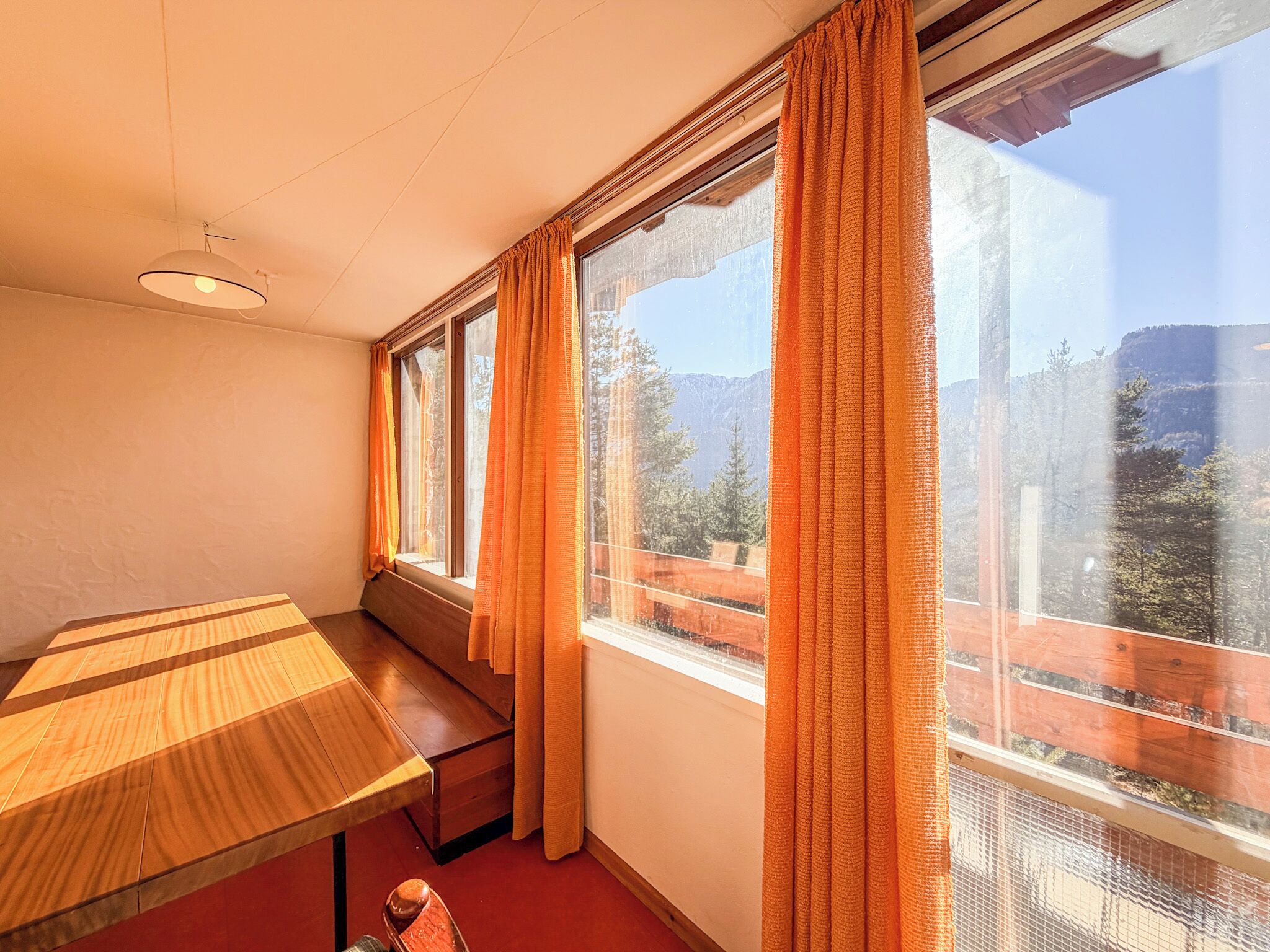 A room with a wooden table and bench, large windows with orange curtains, and a view of a mountainous landscape.