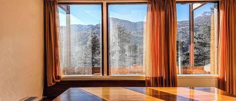 A room with a large wooden table, a bench, and a view of a mountainous landscape through large windows with curtains.