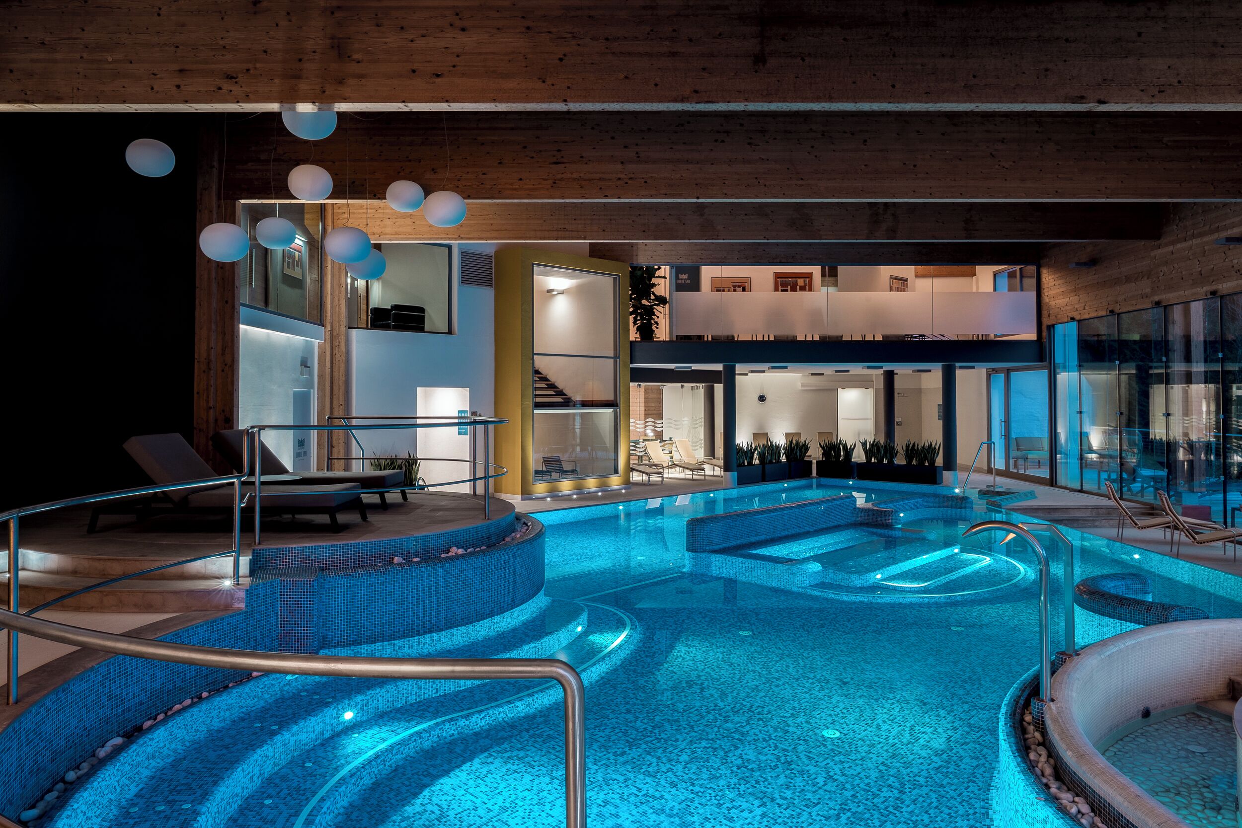A modern indoor swimming pool with clear water, surrounded by lounge chairs and a glass-enclosed area with seating.