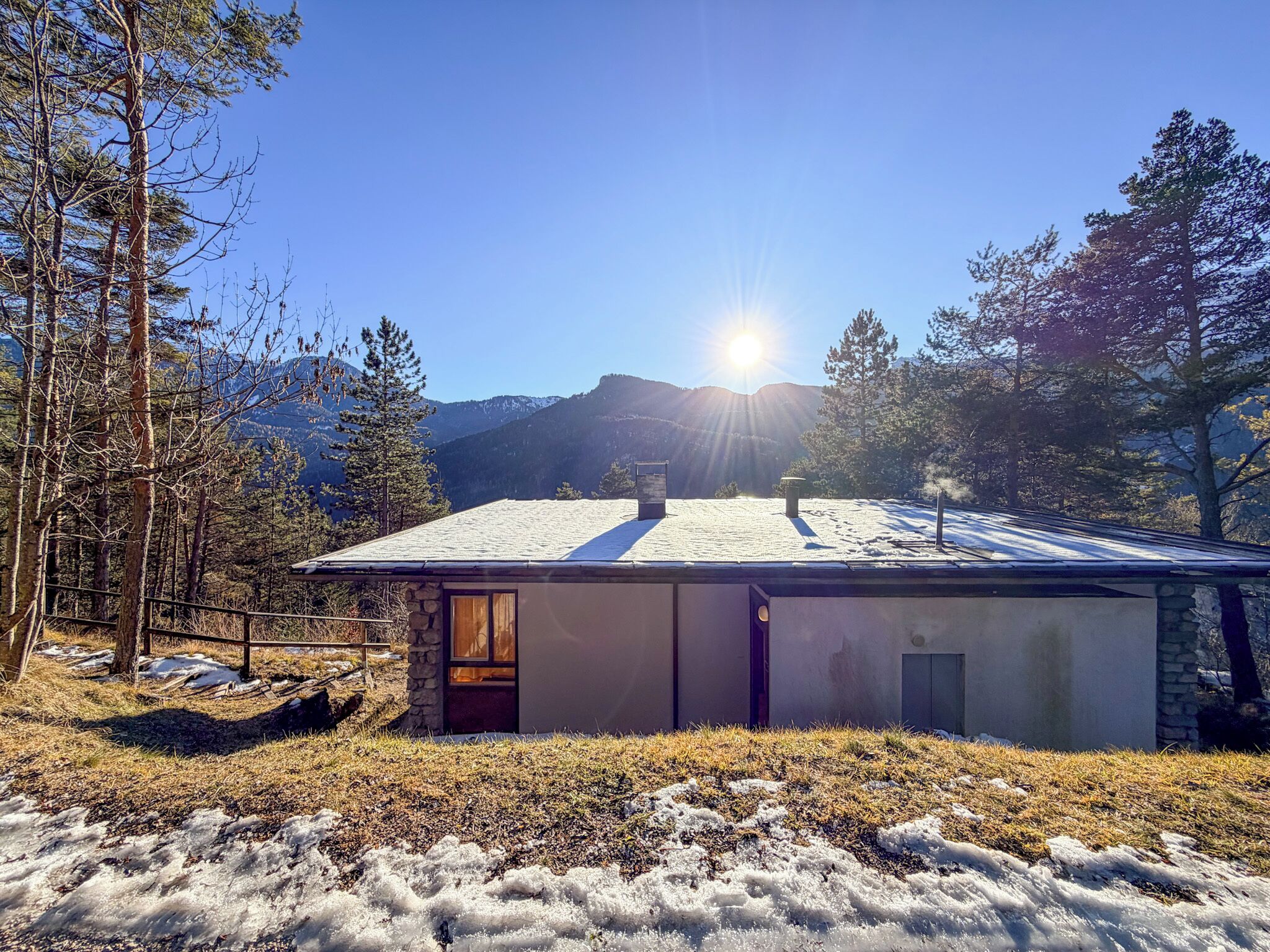 A single-story house with a sloped roof, surrounded by trees and a mountainous backdrop.
