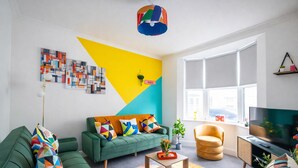 A modern living room with a green sofa, a yellow and blue wall art, a television, and a large window. - Brighton Colour Pop House-Sleeps up to 12 (Brighton and Hove)