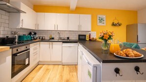 A modern kitchen with white cabinets, a black countertop, and a yellow wall. There is a sink, a microwave, and a dish rack. A vase with flowers and a plate of croissants are on the counter. - Brighton Colour Pop House-Sleeps up to 12 (Brighton and Hove)