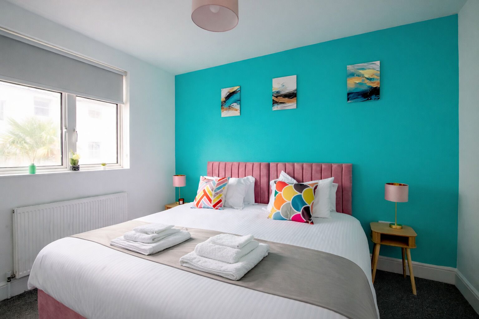 A bedroom with a teal wall, a pink headboard, and three paintings. There are two bedside tables with lamps, a window with a view, and a bed with white linens and colorful pillows.