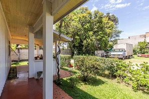 A covered porch with a brick pathway leading to a garden area with various plants and a tree.