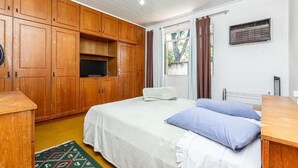 A bedroom with wooden cabinets, a bed with white and blue bedding, a small window with curtains, and a radiator.