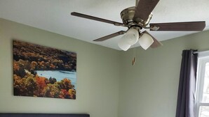 Ein ordentlich bezogenes Bett mit weißen Leinen, einem Deckenventilator und einem Landschaftsbild an der Wand.