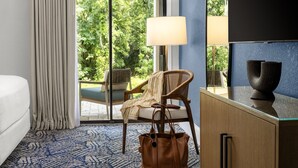 A hotel room with a bed, a chair, a desk, a lamp, a handbag, and a view of greenery through a glass door.