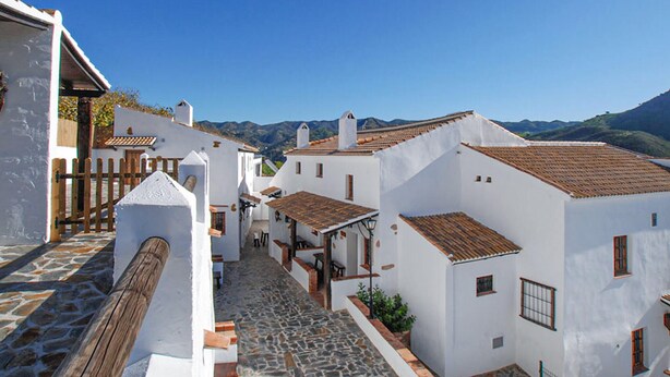 Una calle estrecha con edificios blancos y tejados de teja, que conduce hacia un telón de fondo montañoso.