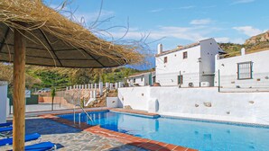 Una piscina con baldosas de piedra y borde azul, rodeada de edificios blancos con balcones y una estructura de techo de paja.