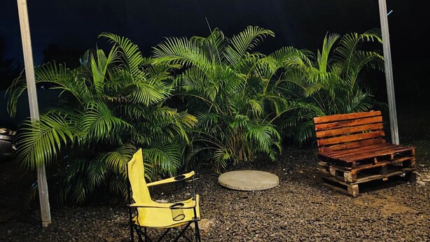 Un espacio al aire libre cubierto con una silla plegable amarilla, un banco de madera y una mesa redonda de piedra.