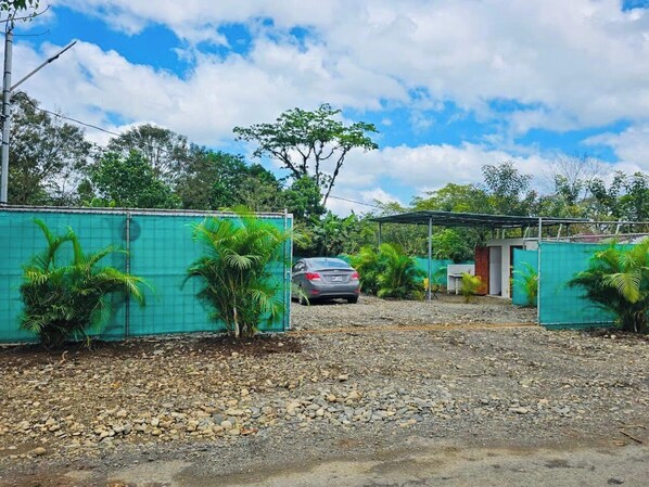 A gravel driveway leading to a car parked in a covered area with green netting on the sides. - Arenal Camping (Penas Blancas)