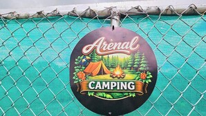 Señal de camping con la palabra 'Arenal' y 'CAMPING' visible, fijada a una cerca de malla metálica.
