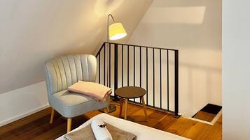 Ein gemütliches Zimmer mit hoher Decke, freiliegenden Holzbalken, einem bequemen Sessel, einem kleinen runden Tisch und einem ordentlich gefalteten Handtuch auf einem weißen Bett.