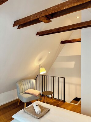 Una habitación acogedora con techo alto, vigas de madera a la vista, un sillón cómodo, una mesita redonda y una toalla blanca doblada con cuidado sobre la cama.