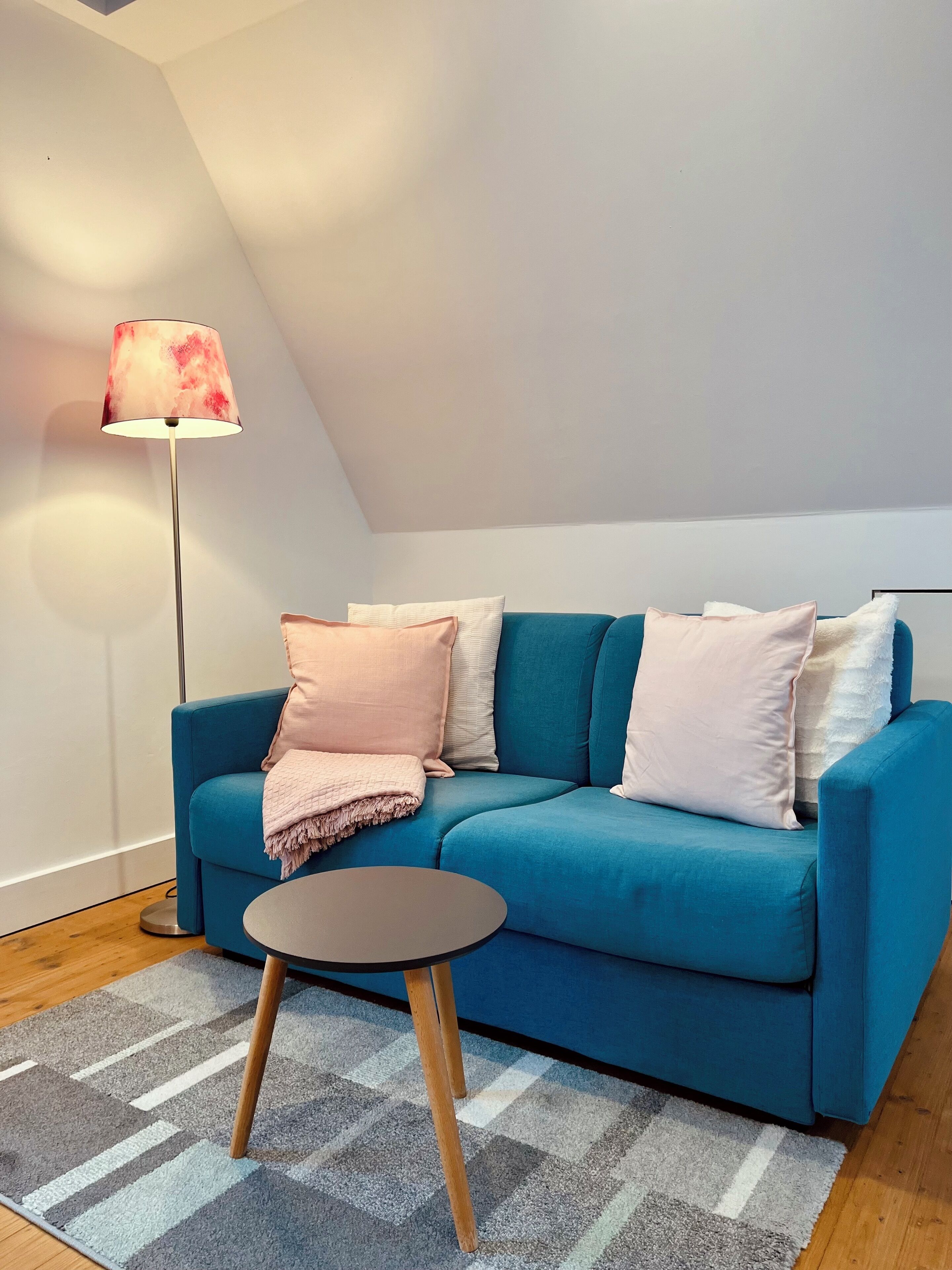Ein modernes Wohnzimmer mit einem blauen Sofa, einem kleinen runden Tisch, einer Stehlampe und einem gemusterten Teppich.