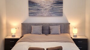 Un lit bien fait, avec deux serviettes enroulées, une tête de lit grise et un tableau représentant un paysage marin au-dessus.