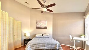 Una camera da letto con un letto, una scrivania, una sedia, una ventola a soffitto, una lampada a parete e un quadro.