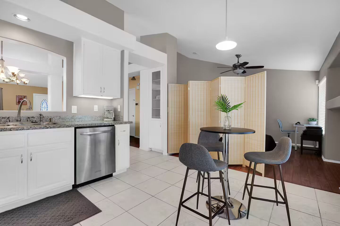 A modern kitchen with white cabinetry, a stainless steel dishwasher, and a granite countertop. There is a dining area with a round table and two bar stools.
