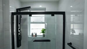 A modern bathroom with a glass shower enclosure, a white toilet, and a window with a plant. - Bright & Modern 3-Bed House ~ Parking Included ~ Near Transit in East Flatbush (Brook)