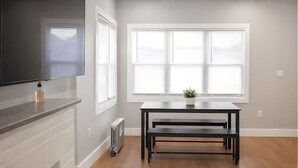 A modern living room with a flat-screen TV, a fireplace, a dining table with benches, and a large window with blinds. - Bright & Modern 3-Bed House ~ Parking Included ~ Near Transit in East Flatbush (Brook)