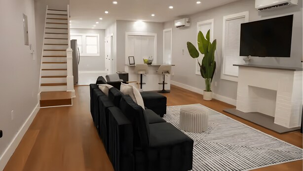 A modern living room with a black sectional sofa, a large TV, and a staircase leading to another room. - Bright & Modern 3-Bed House ~ Parking Included ~ Near Transit in East Flatbush (Brook)