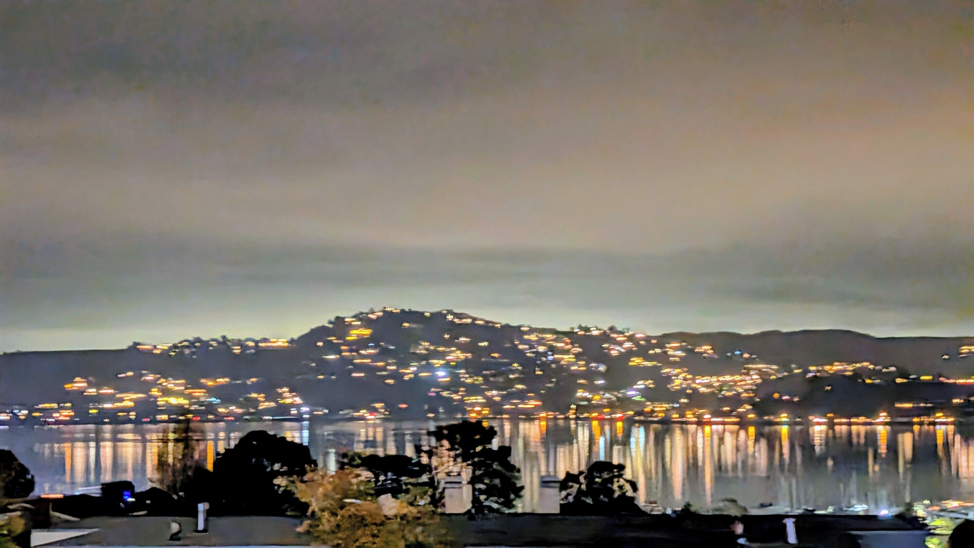 Vista nocturna de un paisaje urbano con edificios iluminados sobre una masa de agua, una colina al fondo y algunos árboles en primer plano.