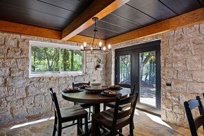 A stone-walled room with a round table and chairs, a chandelier, and a view of trees through large windows.