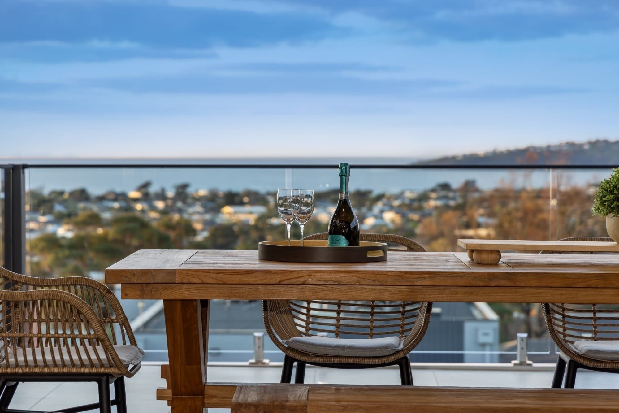 Uma mesa de madeira com duas cadeiras, uma garrafa de champanhe e dois copos numa varanda com vista para a paisagem urbana.