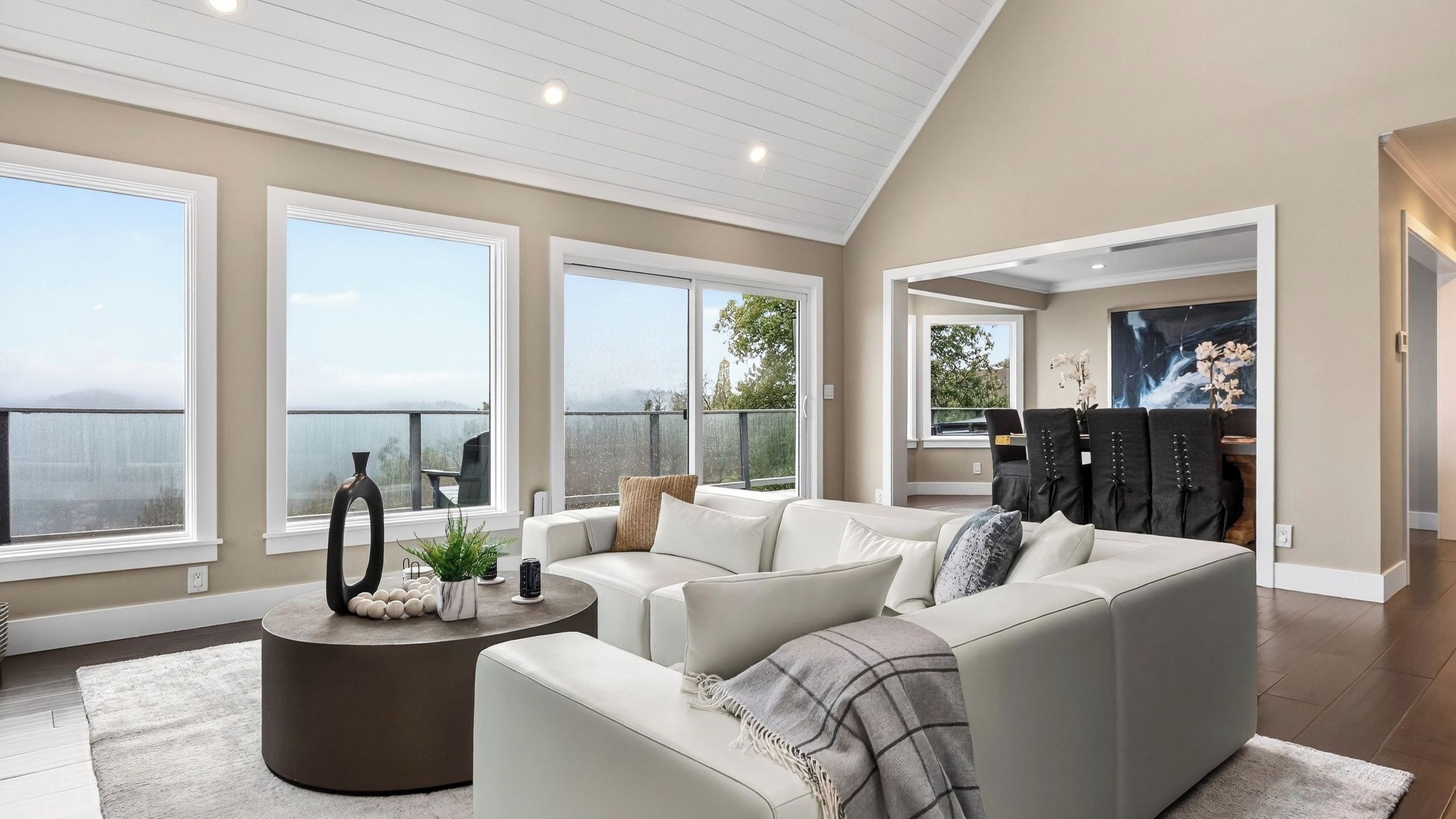 Ein geräumiges Wohnzimmer mit hohen Decken, großen Fenstern mit Blick ins Freie, einem runden Couchtisch und einer Sitzecke mit Sofa und Sesseln.