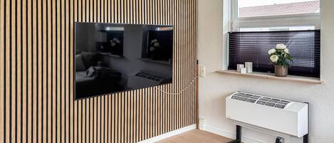 Una stanza con una grande TV a schermo piatto montata su una parete di listelli in legno, una finestra con persiane, un piccolo ripiano con oggetti decorativi e un condizionatore d'aria bianco.