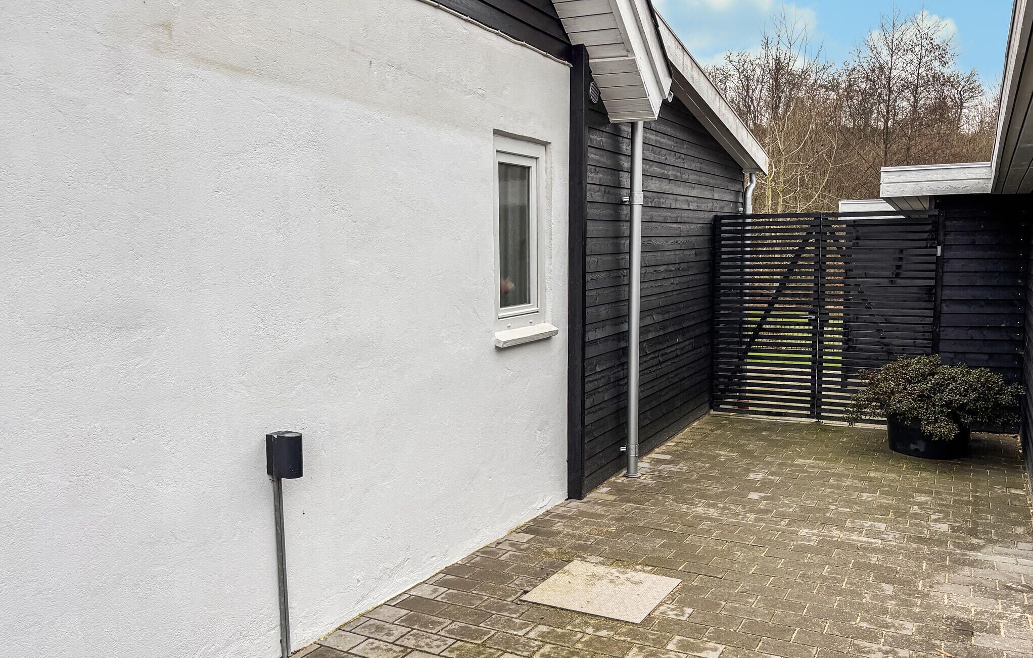 Un estrecho pasillo exterior con una pared blanca a un lado y una valla negra al otro. Se observa una ventana con marco blanco y un pequeño estante debajo. Un tubo de metal recorre la pared, y una planta en maceta se ve a lo lejos.