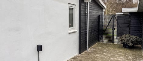 Un estrecho pasillo exterior con una pared blanca a un lado y una valla negra al otro. Se observa una ventana con marco blanco y un pequeño estante debajo. Un tubo de metal recorre la pared, y una planta en maceta se ve a lo lejos.