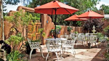 An outdoor patio with white metal chairs and tables, red umbrellas, and a rustic stone wall.