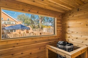 Eine Holzdiele mit großem Fenster, einer Sauna und Blick auf einen Hinterhof mit Zaun und Bäumen.