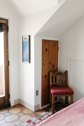 Una habitación con una puerta de madera, una silla roja, un tapete estampado y una lámpara pequeña fijada en la pared.
