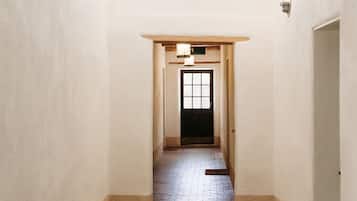 Un couloir avec un sol en briques, des murs blancs et un ventilateur de plafond. Une porte se trouve au bout du couloir, et un luminaire est installé au-dessus.