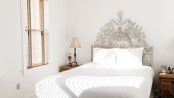 A bedroom with a white bed, a decorative headboard, a bedside lamp, and a window with blinds.