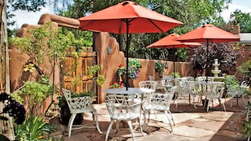 An outdoor patio with white metal chairs and tables, red umbrellas, and a rustic stone wall.