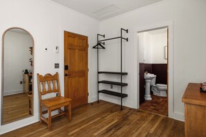 Une pièce avec une porte en bois, un miroir, une chaise en bois, un présentoir en métal noir et des toilettes visibles à travers une porte entrouverte.