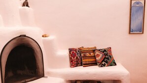 Una chimenea blanca con una reja, una pequeña cesta debajo y tres cojines coloridos sobre un estante blanco frente a una pared lisa.