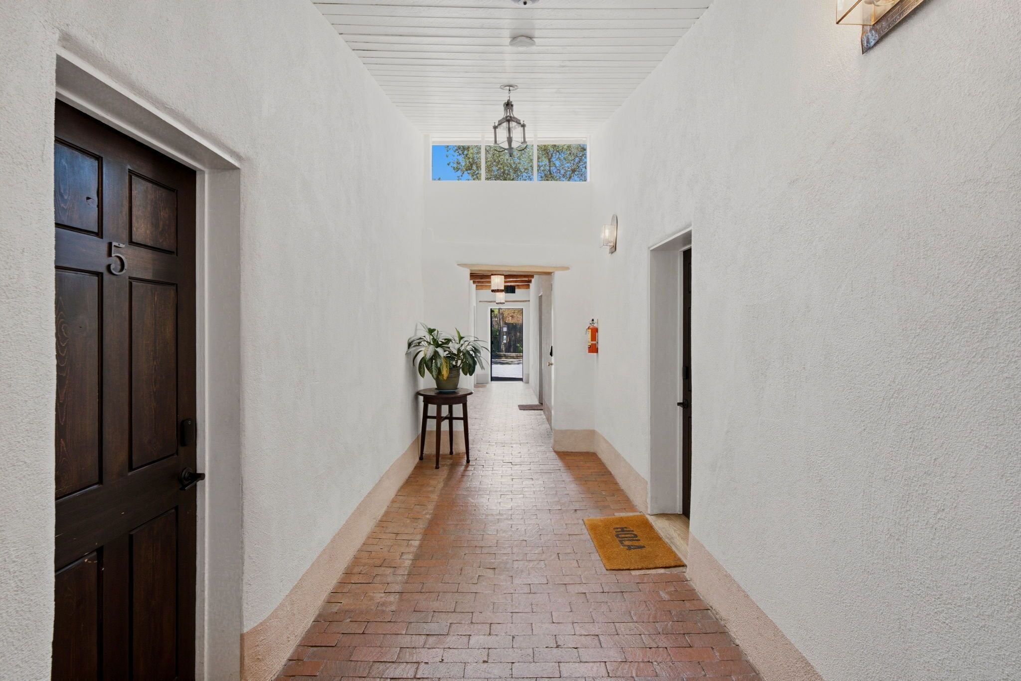 A hallway with a brick floor, white walls, a wooden door with a number, a potted plant, and a door with a fire extinguisher.