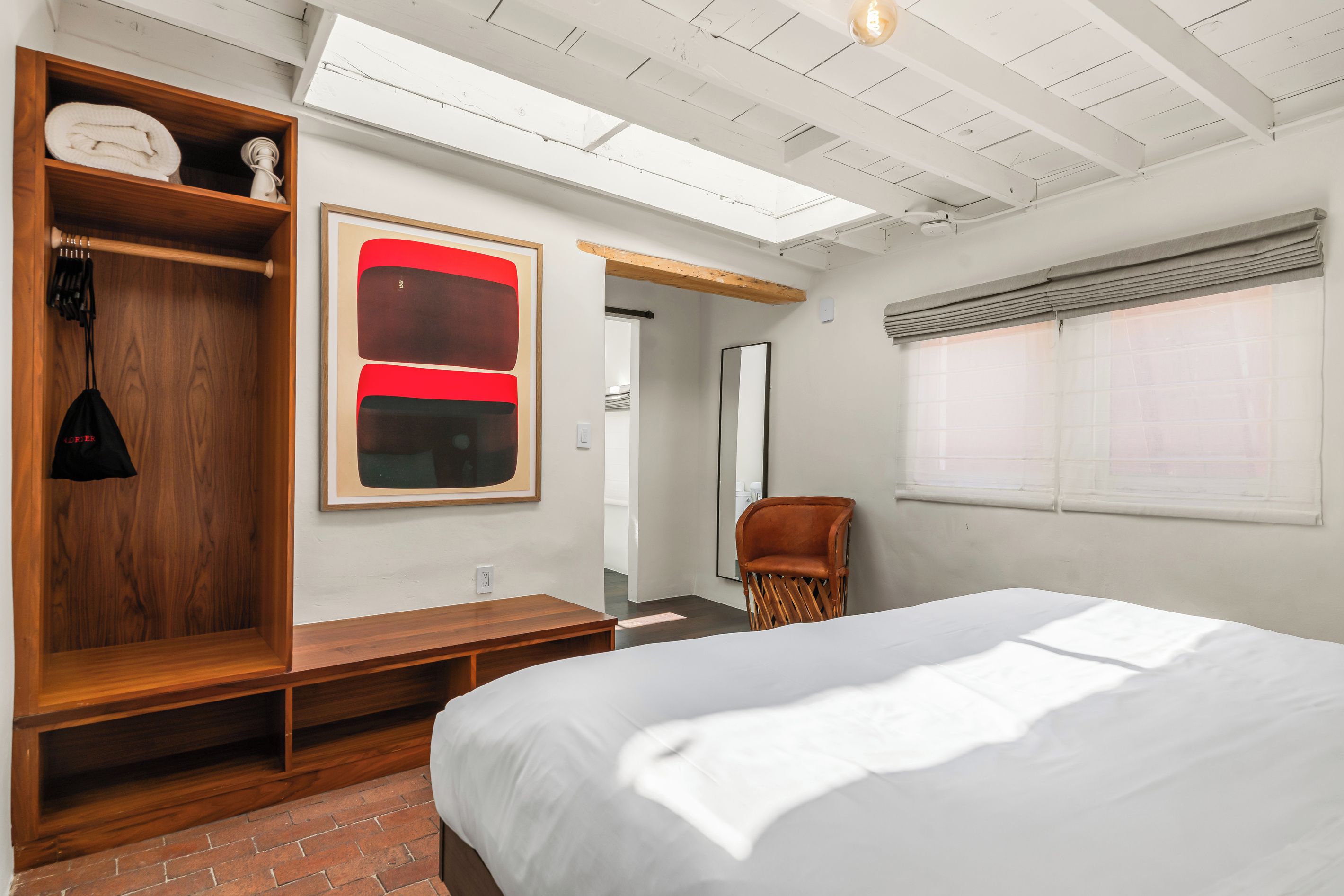 Un dormitorio con cama, un banco de madera, un armario, un espejo y un cuadro abstracto rojo.