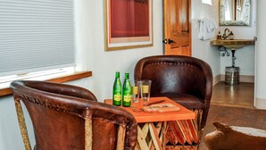 Een kamer met een houten plafond, een raam met luiken, een bruine leren fauteuil, een houten tafel met flessen en glazen, en een spiegel boven een wastafel.