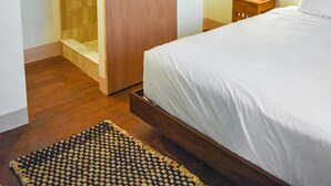 Een slaapkamer met een houten plafond, een bed met wit beddengoed, een houten nachtkastje, een klein tafeltje, een hanglamp en een houten deur naar een douchecel.
