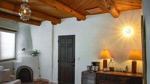 Een gezellige woonkamer met een open haard, een houten plafond en een deur. Op een houten tafel staan twee lampen en er is een klein kastje met lades.