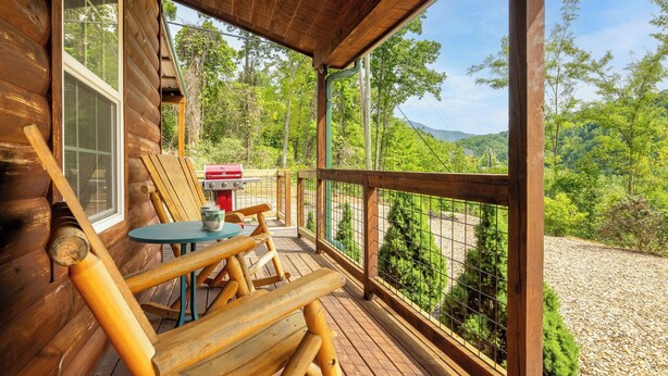 A wooden deck with a table and chairs, a grill, and a view of a forested area.